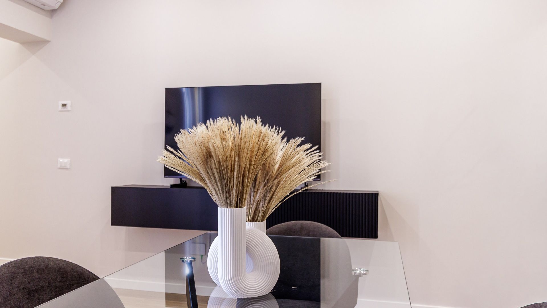 Un salon moderne avec une table basse en verre, un vase blanc contenant des plantes séchées, un téléviseur à écran plat fixé au mur et un canapé de couleur foncée.