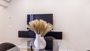 Ein modernes Wohnzimmer mit einem Glastisch, einer weißen Vase mit Trockenpflanzen, einem an der Wand befestigten Flachbildfernseher und einem dunkelgrauen Sofa.