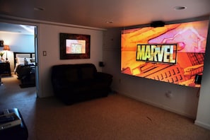 A living room with a large wall-mounted TV displaying a Marvel movie poster, a couch, and a door leading to another room. - Cozy One Private Room and Bath in Pentagon City House (Arlington)
