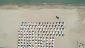 Vista aerea di una spiaggia con file di ombrelloni e lettini, alcune persone e una persona che cammina sulla sabbia.