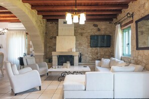 A spacious living room with a stone fireplace, white sofas, and a flat-screen TV mounted on the wall. - Villa Pinelopi - 350 meters away from sandy beach! (Rethymno)