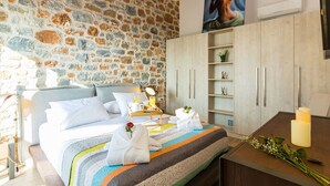 A bedroom with a stone wall, a bed with a striped blanket, a bedside table with a lamp, a wardrobe, and a small shelf with flowers. - Beautiful sea views, Close to Beautiful Beaches of South Crete (Rethymno)