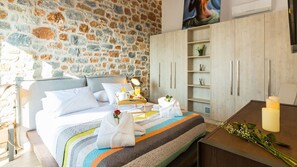 Um quarto com parede de pedra, cama com manta listrada, mesa de cabeceira com abajur, guarda-roupa e uma pequena prateleira com flores.