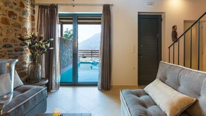 Un salon avec un mur en pierre, un canapé, une table basse et une vue sur une piscine et des montagnes, visible à travers une porte vitrée.