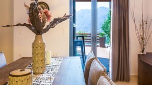 Un coin repas avec une table dressée pour deux personnes, un vase rempli de fleurs et une vue sur les montagnes, visible à travers une porte vitrée.