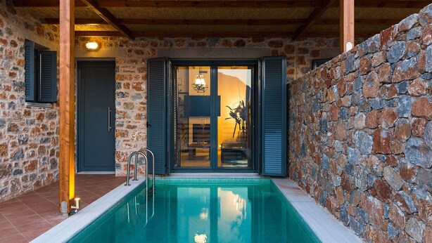 Un espace piscine entouré de murs en pierre, avec une piscine à eau claire, des poutres en bois et une vue sur une pièce où se trouvent un lit et une table.