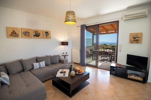 Ein modernes Wohnzimmer mit einer Couch, Couchtisch und einem TV-Schrank. An den Wänden befinden sich Gemälde und es gibt einen Balkon mit Blick.
