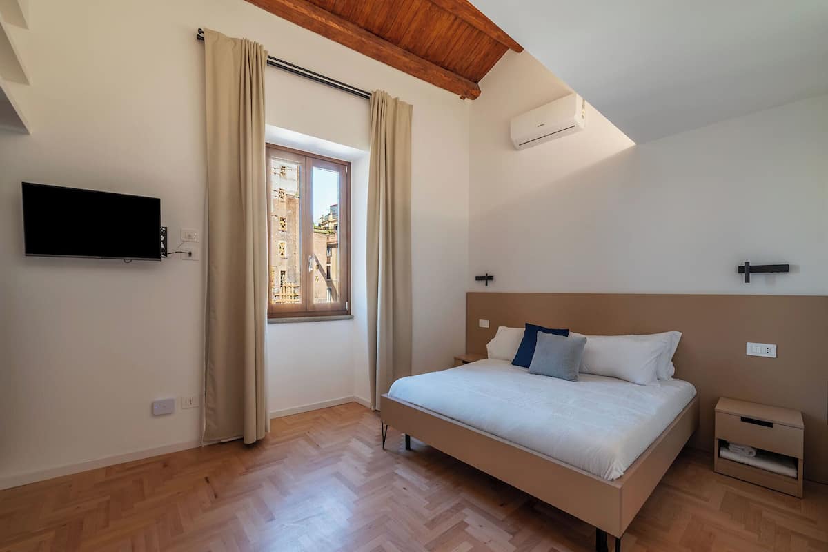 Una camera da letto con un letto, una TV, una finestra con tende, un pavimento in legno e un condizionatore d'aria.