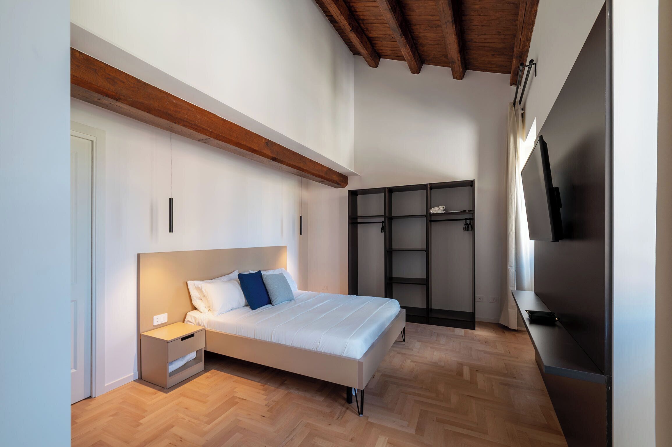 Um quarto com cama, mesa de cabeceira, guarda-roupa e televisão.