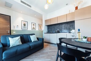 A modern living room with a dark blue sofa, a dining table with chairs, and a kitchen area with cabinets and a countertop. - Ciclamino - Modern apartment with private parking. Lake view (Bellano)