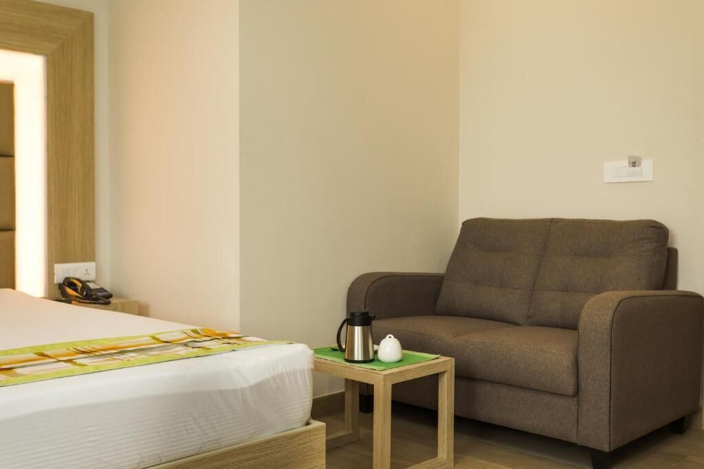 A hotel room with a bed, a brown armchair, a small wooden table with a kettle and a cup, and a wall-mounted telephone.