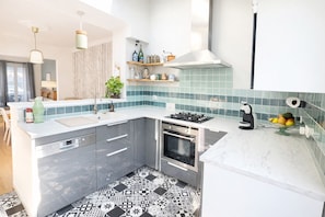 Een moderne keuken met een ingebouwde oven, een gasfornuis en een spoelbak. Op het aanrecht staat een fruitschaal en op de vensterbank een plant.
