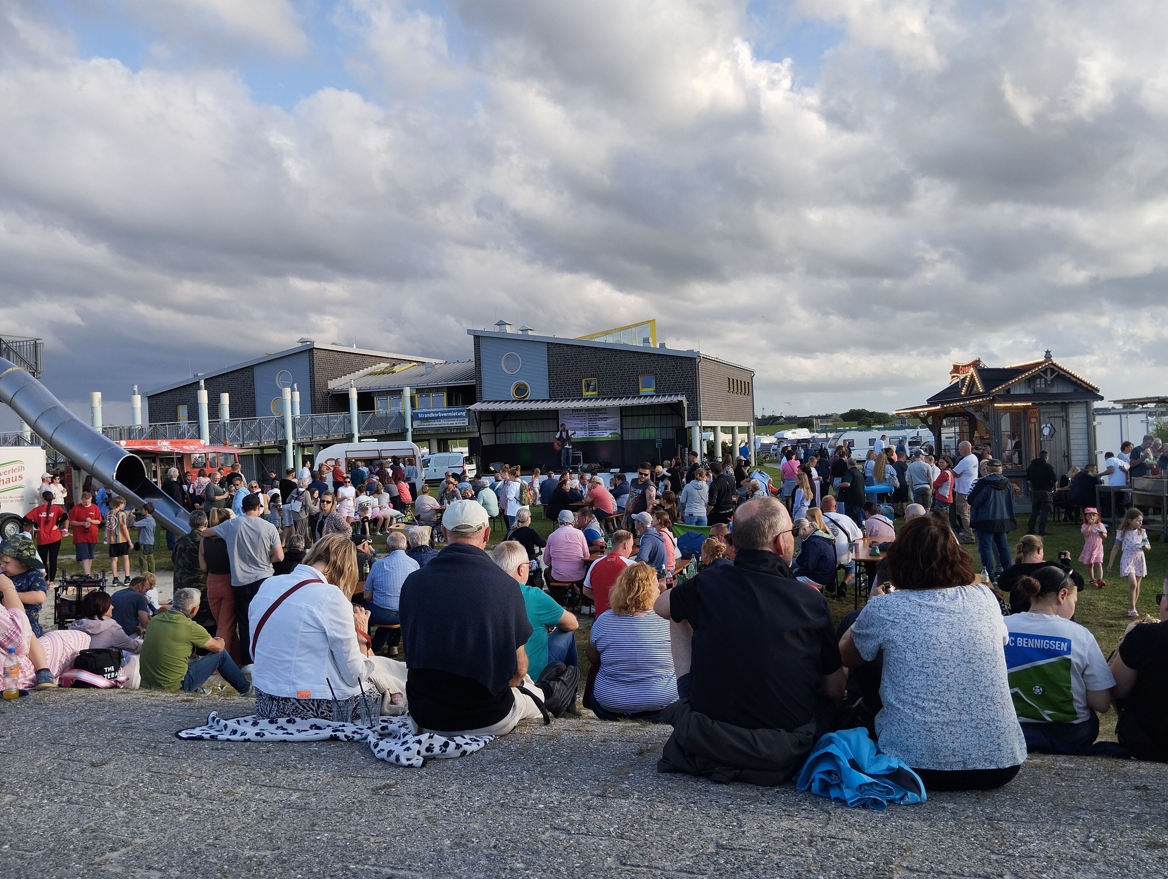 A crowded outdoor event with people sitting on the ground, a stage, and buildings in the background.
