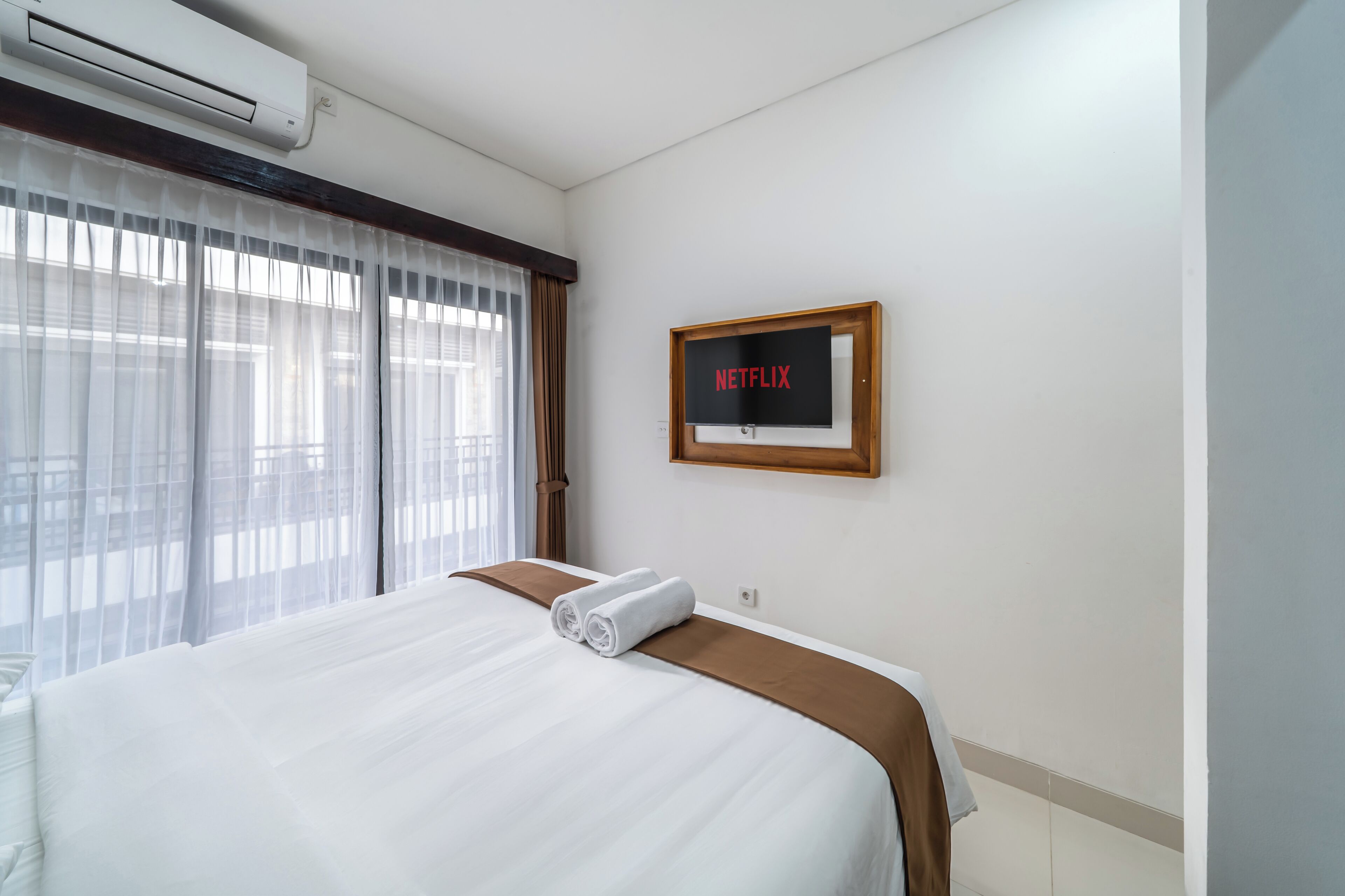 A hotel room with a bed, a wall-mounted TV displaying 'NETFLIX,' and a window with sheer curtains.