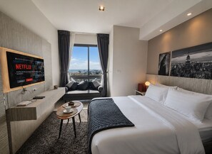 A hotel room with a large bed, a TV mounted on the wall, a sofa, a small table with a cup and saucer, and a view of the city through the window. - Sea view condo @ Unixx south pattaya close to walking street and beach Pattaya (ตำบล หนองปรือ)