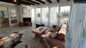 Ein geräumiges Wohnzimmer mit einer großen Couch, einem Couchtisch und einem Fernseher. Eine Glastür mit Schieberegner führt auf einen Balkon mit Blick ins Freie.