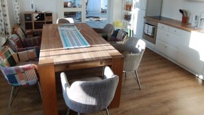 Ein geräumiges Wohnzimmer mit Holztisch, bunten Stühlen, einer Couch und einer Küchenzeile mit weißen Schränken und einem Kühlschrank.