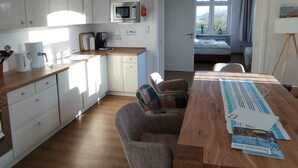 Eine moderne Küche mit weißen Schränken, einer Mikrowelle und einer Kaffeemaschine. Ein Essbereich mit Holztisch und Stühlen, und durch eine offene Tür ist ein Schlafzimmer zu sehen.