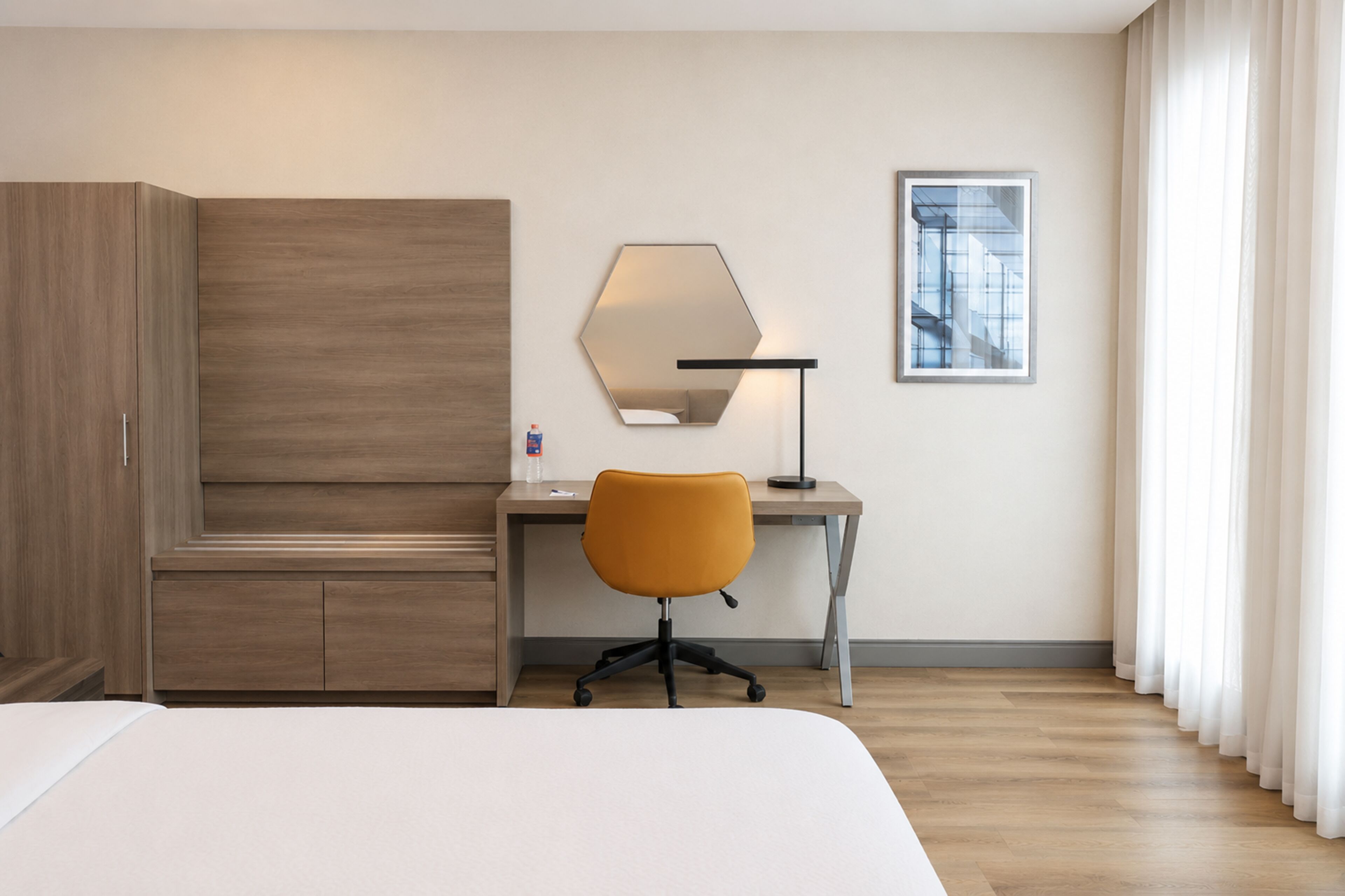 A hotel room with a bed, a desk, an orange office chair, a large mirror, and a framed picture on the wall.