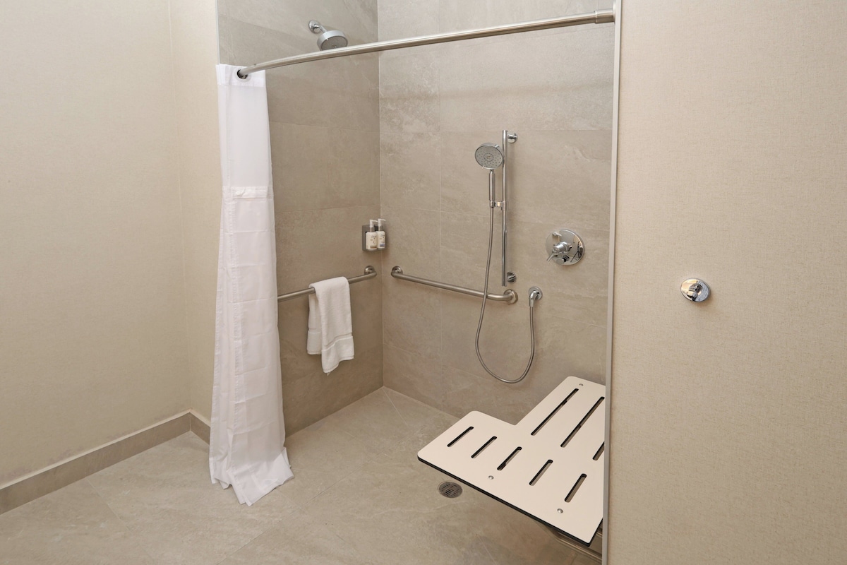 A modern shower area with a glass door, a white shower curtain, a handheld showerhead, a handheld shower, and a white grab bar.