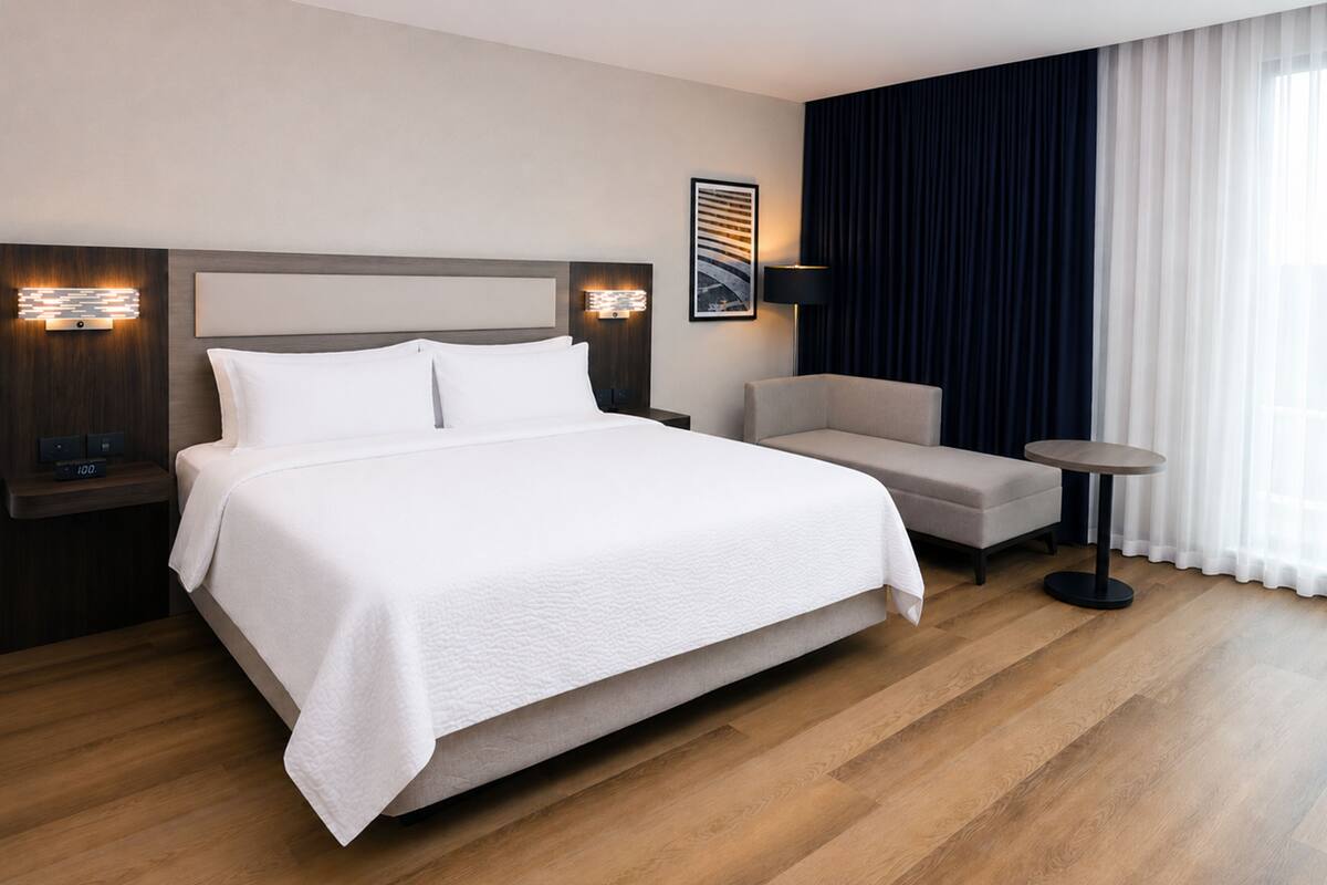 A hotel room with a large bed, a bedside table, a chair, a small round table, and a window with curtains.