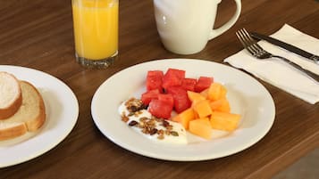 Un desayuno con una fuente de sandía, melón, yogur y granola, un vaso de jugo de naranja y una taza de café sobre una mesa de madera.