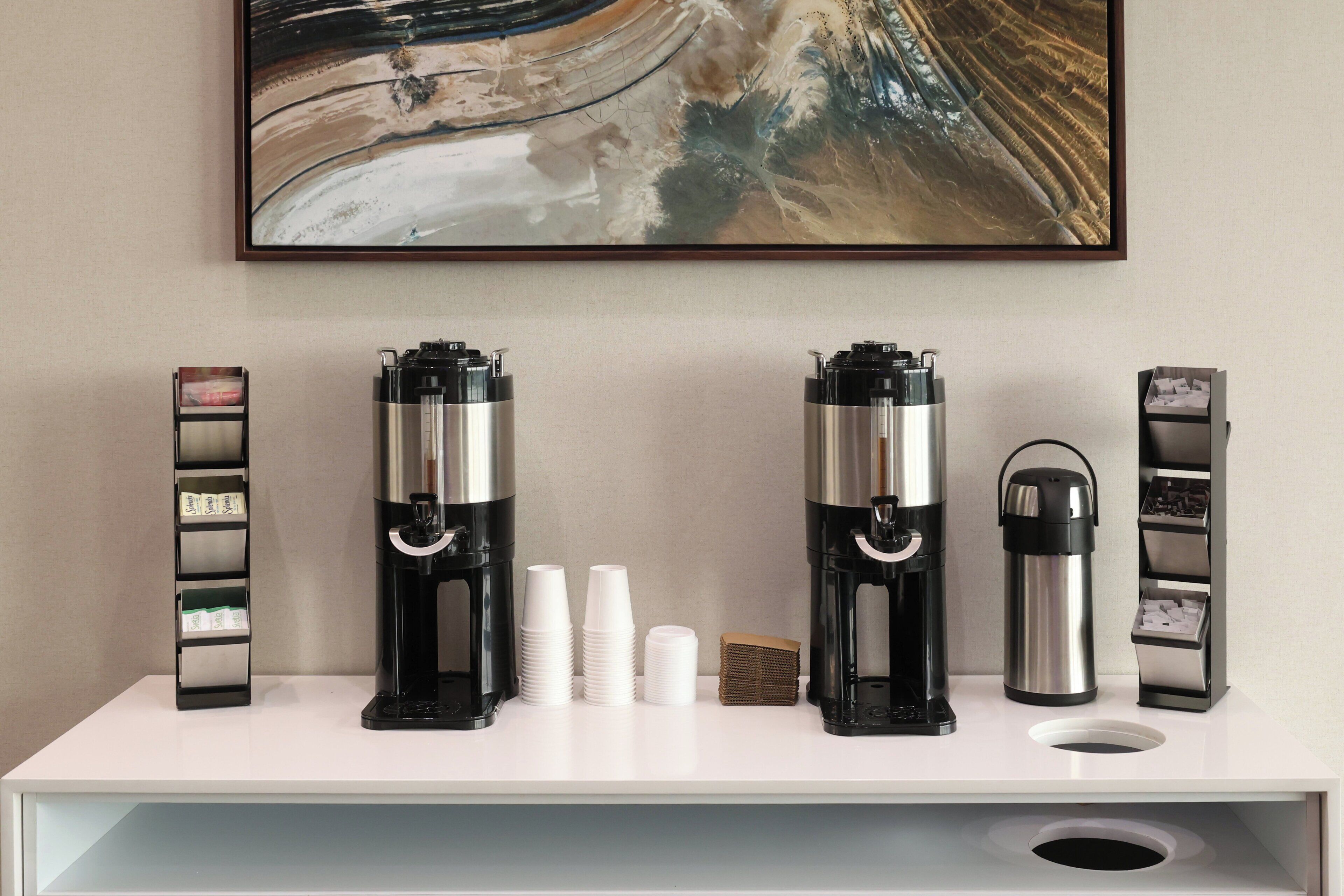 A coffee station with two espresso machines, disposable cups, a thermos, and a cash register on a white counter.