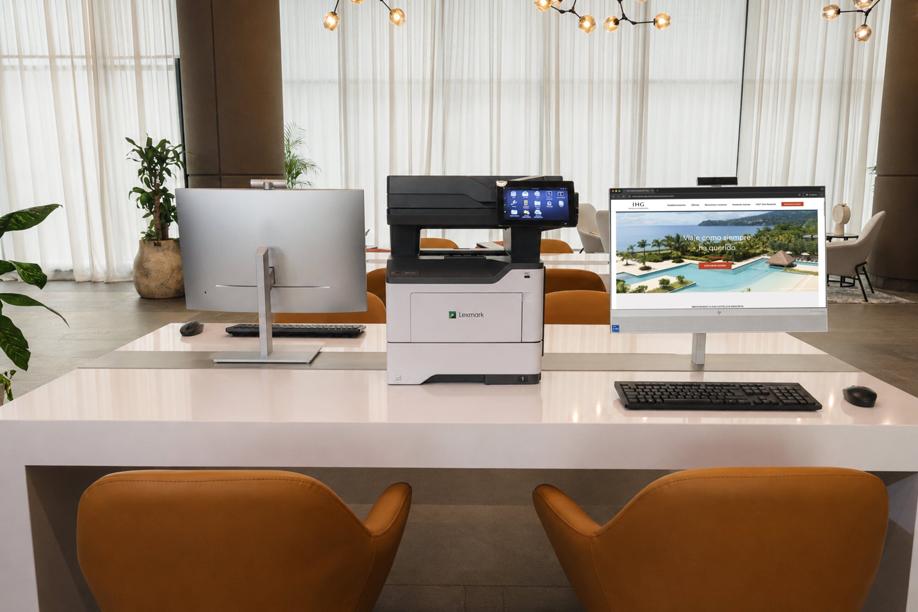 Una oficina moderna con una impresora Lexmark, una computadora de escritorio y un monitor que muestra una imagen de un resort tropical.