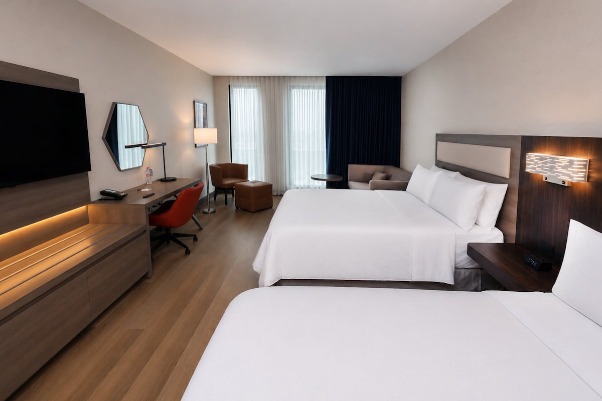 A hotel room with two beds, a desk, a chair, a TV, and a lamp.