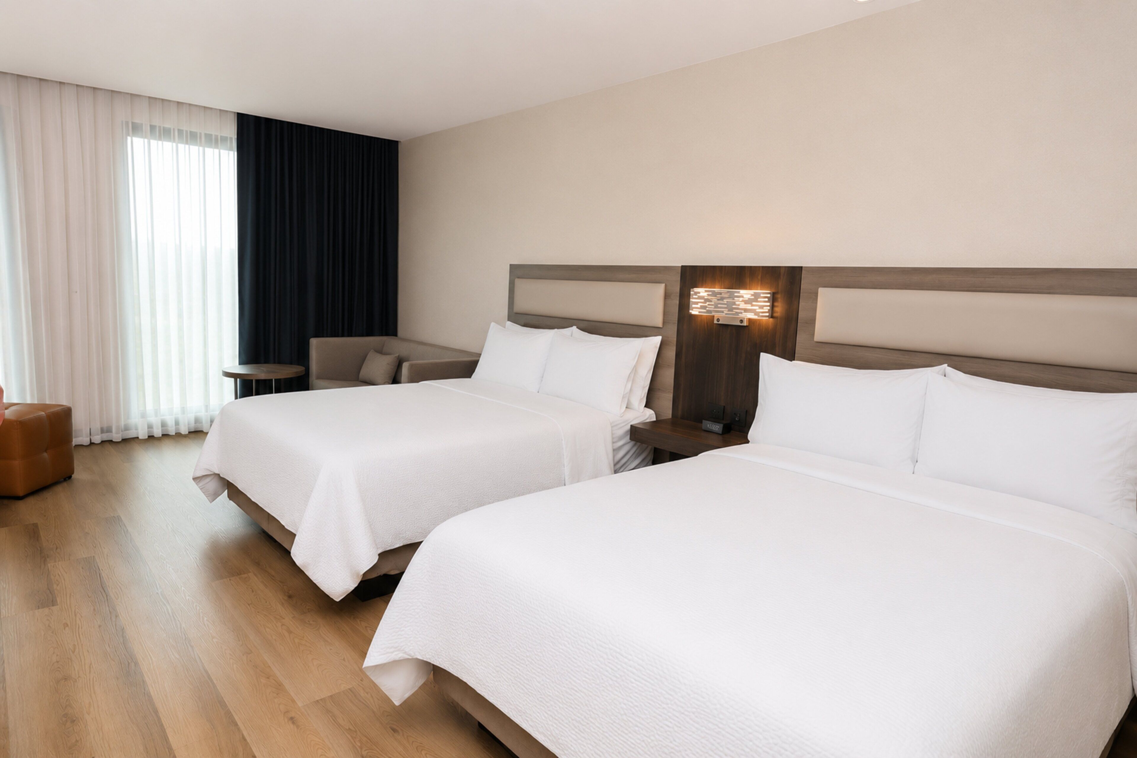 Ein Hotelzimmer mit zwei Betten, einem Nachttisch, einer Lampe, einem Sessel und einem Fenster mit Vorhängen.