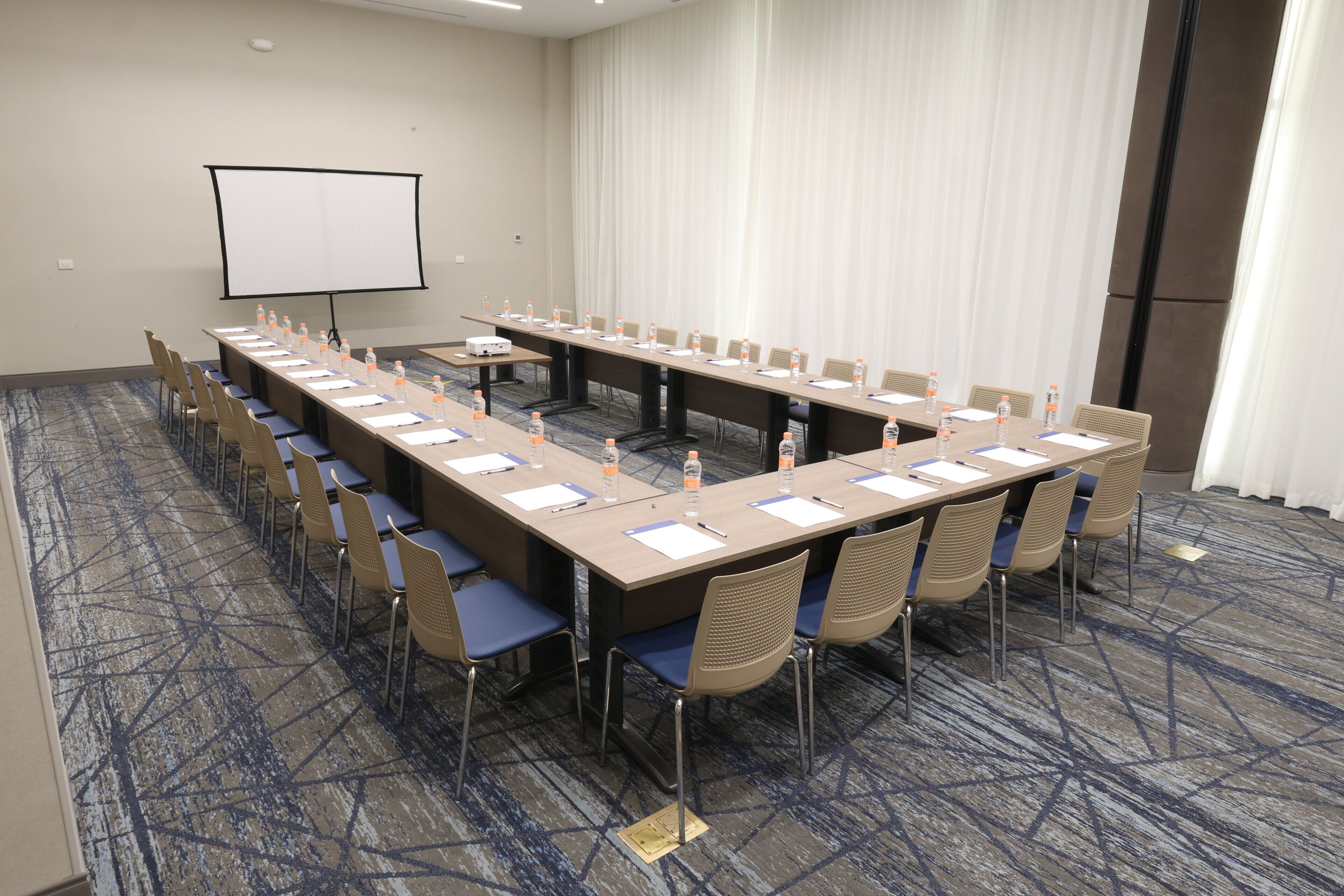 Un salón de conferencias con una larga mesa, sillas, una pantalla de proyector y piso alfombrado.