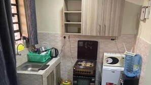Cocina compacta con estufa a gas, microondas y dispensador de agua. Hay una pequeña encimera con un lavamanos y una cesta verde. La habitación tiene un sofá gris con cojines amarillos.