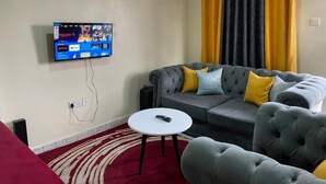 Una sala de estar con un sofá gris, cojines amarillos, un televisor de pantalla plana en la pared, una mesa de centro blanca y una alfombra roja.