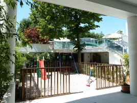 Un'area giochi con scivolo e altalene, circondata da una recinzione in legno, e un edificio con un balcone in vetro sullo sfondo.