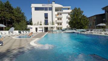 A swimming pool with lounge chairs and umbrellas, surrounded by a building with balconies and a clear sky.