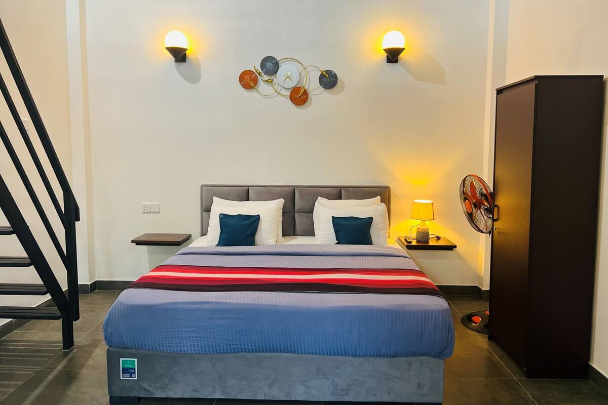 Habitación de hotel con una cama, dos lámparas de pared, un adorno en la pared, una mesita de noche, una lámpara y un armario.