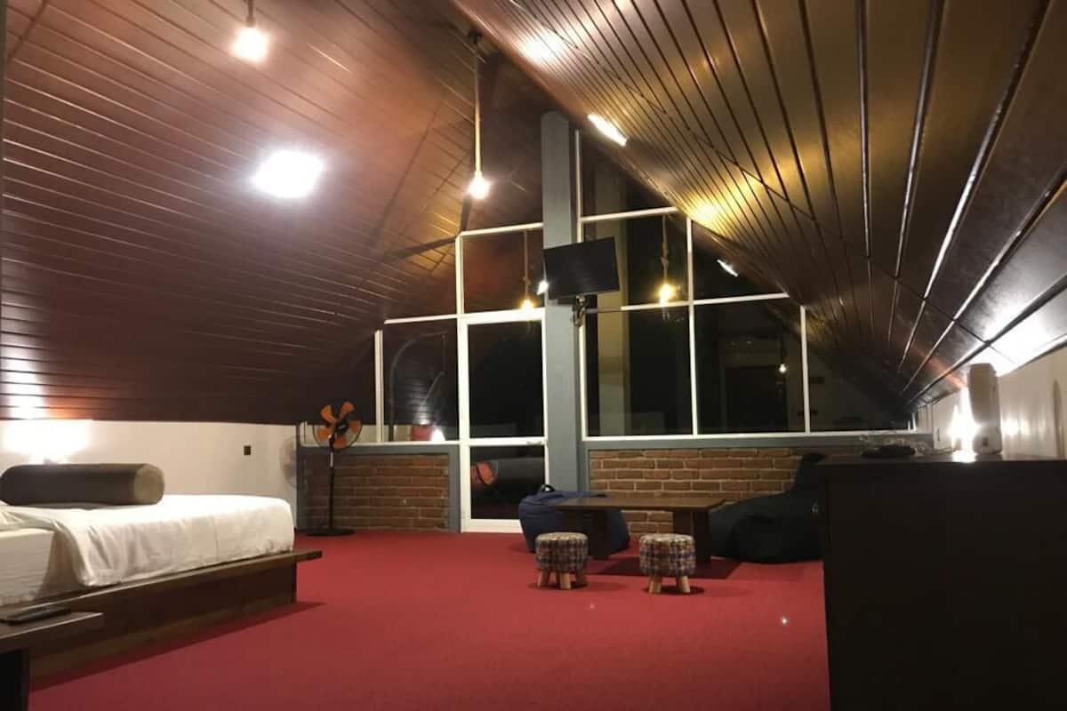 Una habitación con alfombra roja, una cama, dos otomanas escocesas, un ventanal grande y una pared de ladrillo.