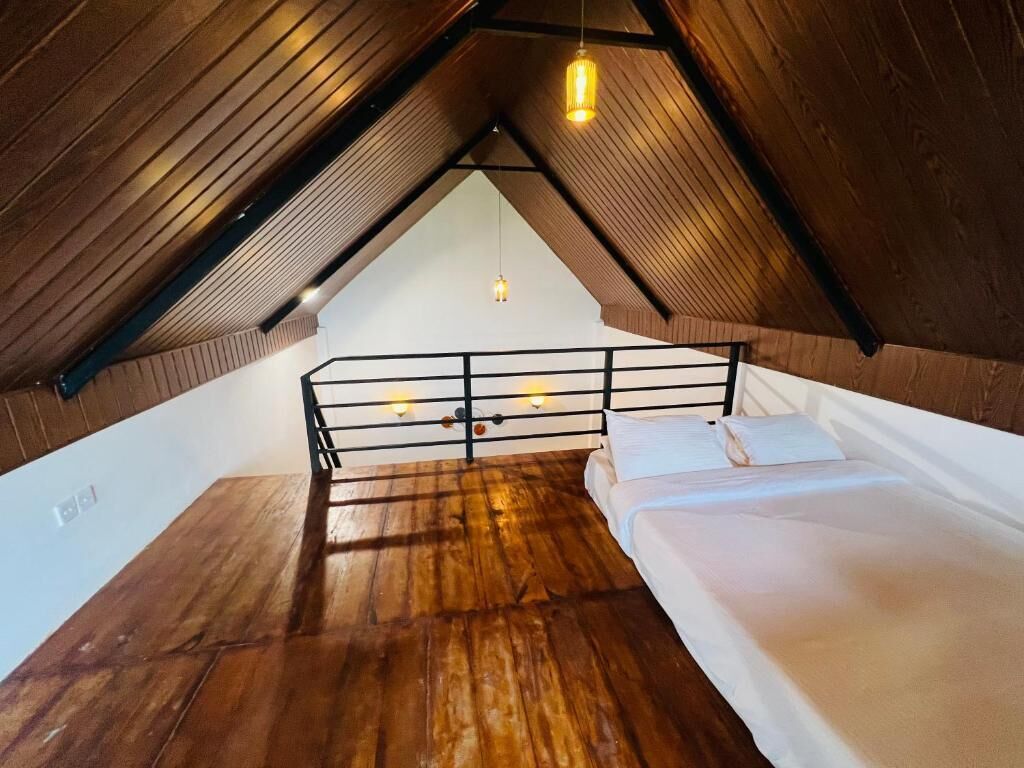 Ein Zimmer mit schrägen Wänden, Holzboden, einem Bett mit weißer Bettwäsche, einem schwarzen Metallgeländer und Pendelleuchten.