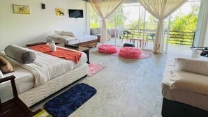 A spacious room with a bed, a sofa, and a TV mounted on the wall. There are large windows with curtains, a ceiling fan, and a view of greenery outside.