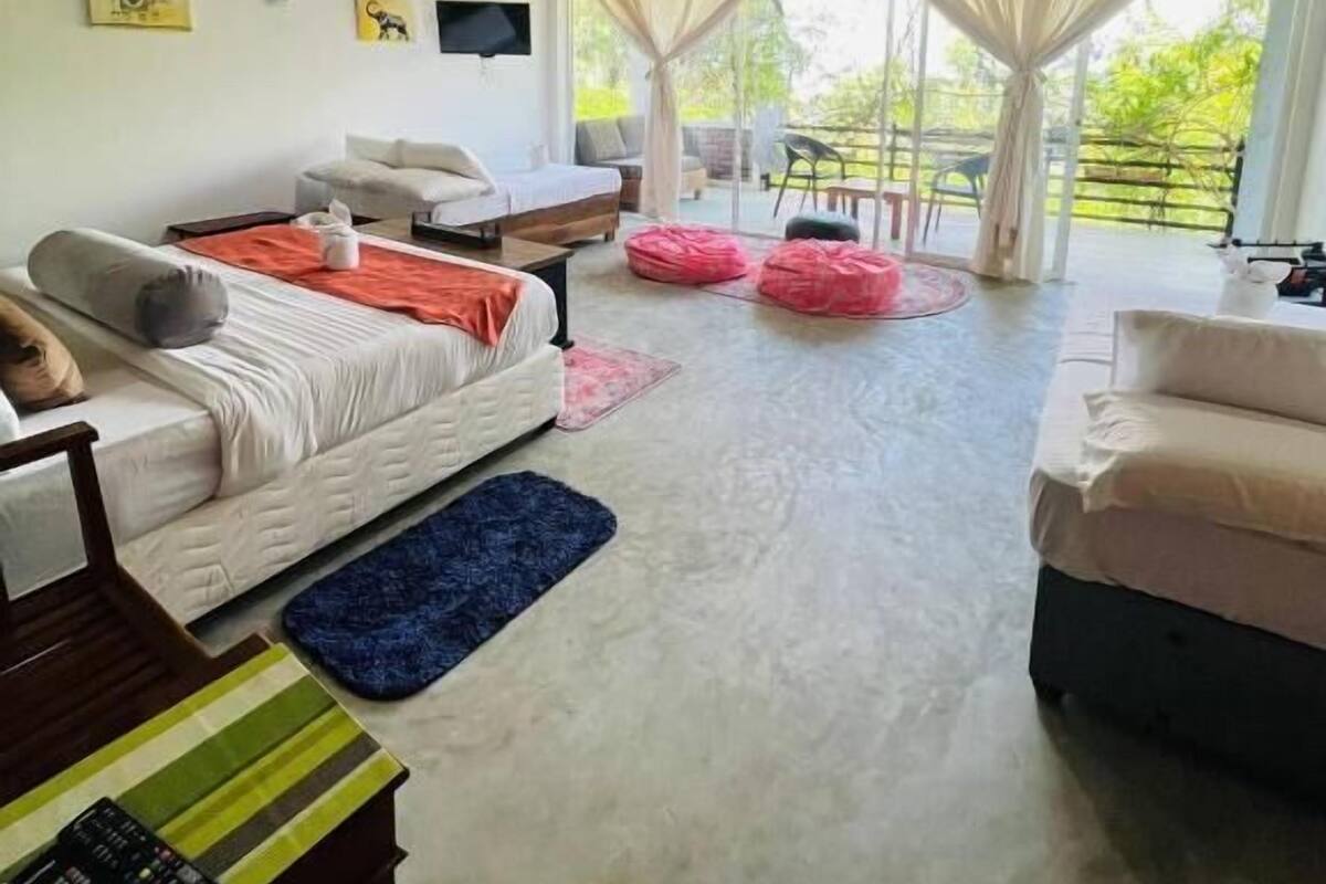 Habitación amplia con cama, sofá y televisor montado en la pared. Cuenta con ventanas grandes con cortinas, ventilador de techo y vistas al exterior con vegetación.