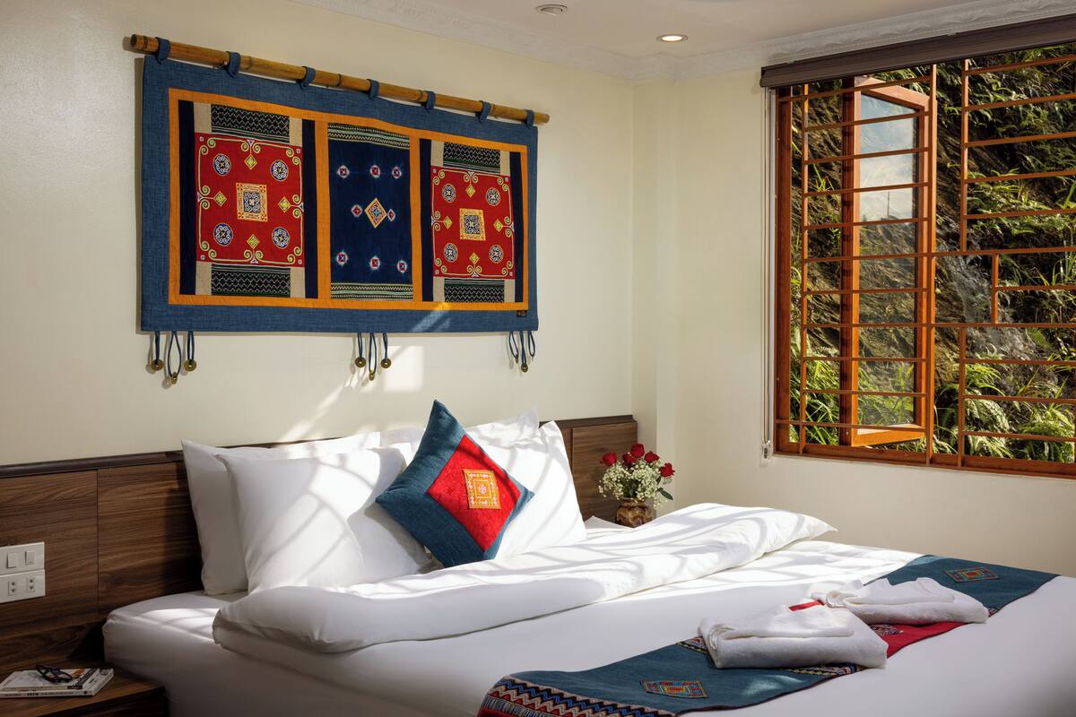 Un lit bien fait, avec du linge de lit blanc et des coussins décoratifs, une décoration murale au-dessus, et une fenêtre donnant sur un paysage verdoyant.