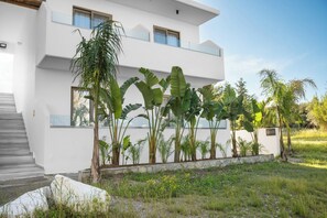 Un edificio moderno de dos pisos con fachada blanca, una escalera que conduce a la entrada y una variedad de plantas tropicales en primer plano.