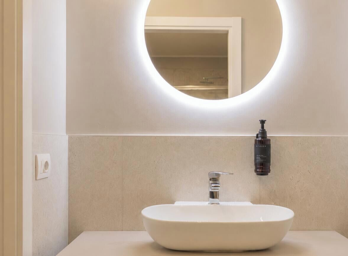Un baño con un espejo redondo, un lavabo blanco y una botella de jabón de manos en la pared.