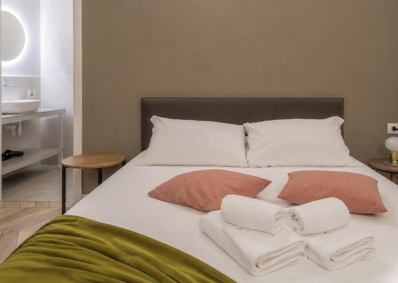 Una habitación de hotel con cama, almohadas, una mesita de noche, una lámpara y un espejo.