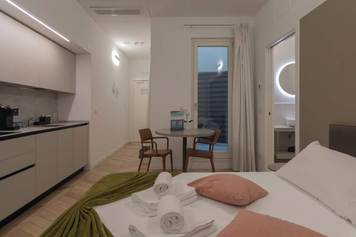 A hotel room with a bed, a small kitchenette, a dining area, and a view of the sky through a window.