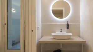 Une salle de bain avec un lavabo blanc, un miroir rond avec un éclairage intégré, un distributeur de serviettes et un porte-rouleau de papier toilette.