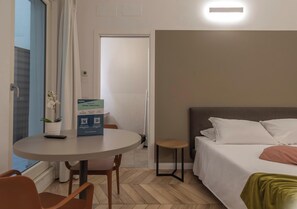 Ein Hotelzimmer mit einem Bett, einem runden Tisch mit Stühlen, einem kleinen Beistelltisch, einer Topfpflanze und einer Tür zu einem anderen Raum.