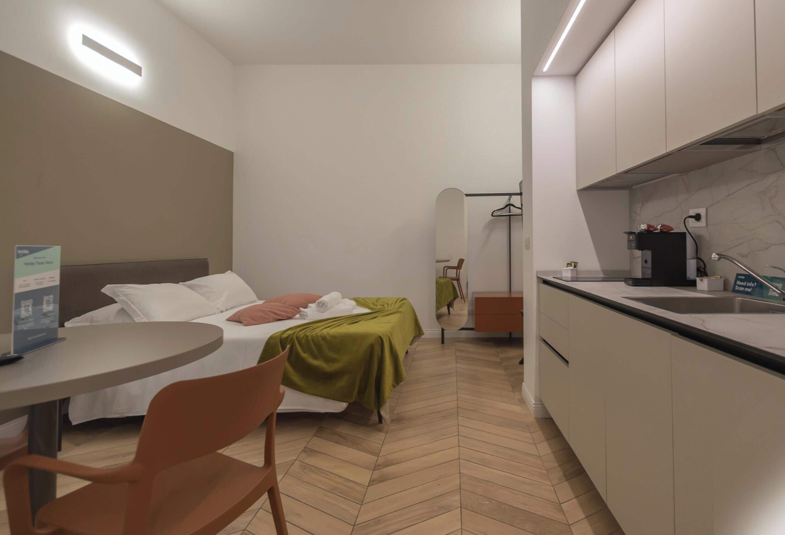 A hotel room with a bed, a table, a chair, a mirror, a dresser, and a kitchenette.