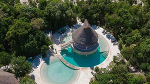 Luftaufnahme eines Resorts mit einem zentralen, kreisförmigen Pool, umgeben von üppiger Vegetation und einem Bauwerk mit einem Strohdach.