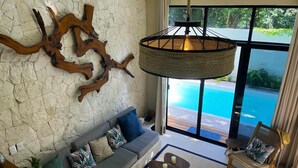 Ein modernes Wohnzimmer mit Steinwand, einer großen Holzskulptur, einem grauen Sofa mit gemusterten Kissen, einer hängenden Leuchte und Blick auf einen Swimmingpool durch große Fenster.